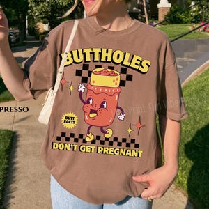 May include: Espresso-colored t-shirt with a cartoon graphic. The graphic features a smiling, anthropomorphic jar with the words "BUTTHOLES DON'T GET PREGNANT" in yellow. The shirt has a relaxed fit.