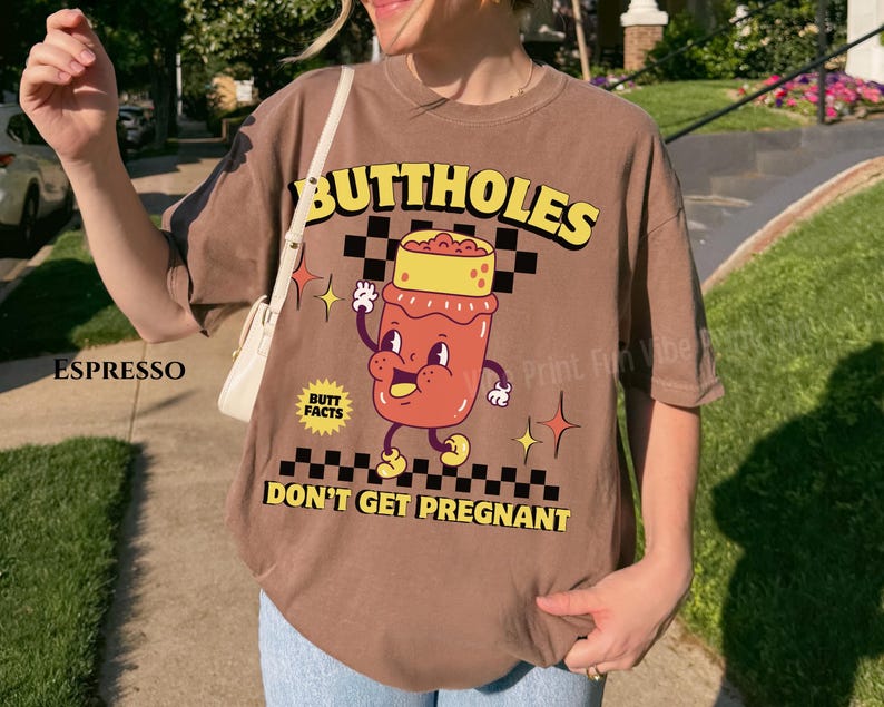 May include: Espresso-colored t-shirt with a cartoon graphic. The graphic features a smiling, anthropomorphic jar with the words "BUTTHOLES DON'T GET PREGNANT" in yellow. The shirt has a relaxed fit.