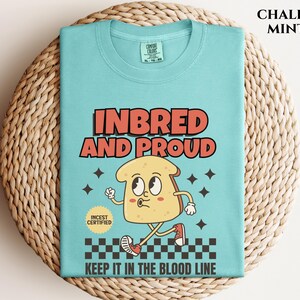 May include: A light blue t-shirt with a cartoon illustration of a slice of bread with a face. The text "Inbred and Proud" is printed in red and white. The text "Keep it in the blood line" is printed in black. The text "Incest Certified" is printed in a circle with a red border.
