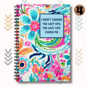 May include: A spiral bound notebook with a white background and a colorful floral pattern. The notebook has a blue cover with the text "I didn't choose the lazy life, the lazy life chose me".