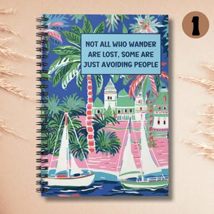 May include: A blue spiral bound notebook with a colorful illustration of palm trees, sailboats, and buildings. The notebook has the text "Not all who wander are lost. Some are just avoiding people." printed on the front cover.