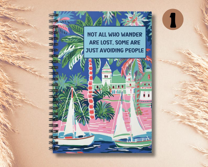 May include: A blue spiral bound notebook with a colorful illustration of palm trees, sailboats, and buildings. The notebook has the text "Not all who wander are lost. Some are just avoiding people." printed on the front cover.