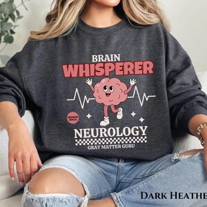 May include: Dark heather grey sweatshirt with a brain graphic and the text "Brain Whisperer Neurology Gray Matter Guru".