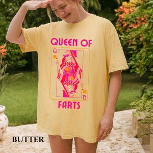 May include: A yellow t-shirt with a graphic of a playing card featuring a queen wearing sunglasses. The text "QUEEN OF FARTS" is printed in pink. The word "BUTTER" is printed at the bottom of the shirt.