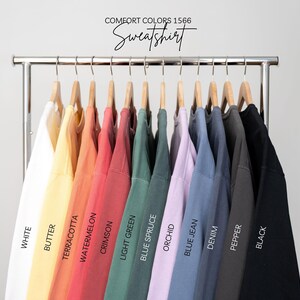 May include: A rack of Comfort Colors 1566 sweatshirts in various colors, including white, butter, terracotta, watermelon, crimson, light green, blue spruce, orchid, blue jean, denim, pepper, and black. The word "Sweatshirt" is written above the shirts.