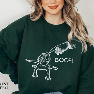 May include: Forest green sweatshirt featuring a white graphic of a lizard being booped on the nose by a finger. The word "BOOP!" is printed next to the lizard. The words "FOREST GREEN" are printed at the bottom left.