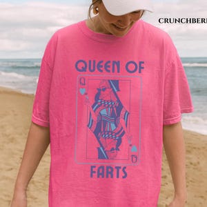 May include: A bright pink t-shirt with the text "QUEEN OF FARTS" in a blue font. The graphic features a playing card design with a woman wearing sunglasses. The shirt is a casual, oversized fit.