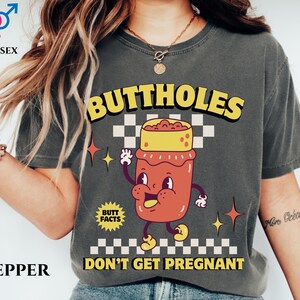 May include: Pepper-colored unisex t-shirt with the text "BUTTHOLES DON'T GET PREGNANT" in yellow and black. The shirt features a cartoon illustration of a jar with a face. The shirt also has the text "BUTT FACTS".