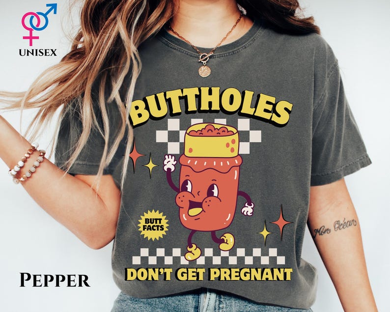 May include: Pepper-colored unisex t-shirt with the text "BUTTHOLES DON'T GET PREGNANT" in yellow and black. The shirt features a cartoon illustration of a jar with a face. The shirt also has the text "BUTT FACTS".