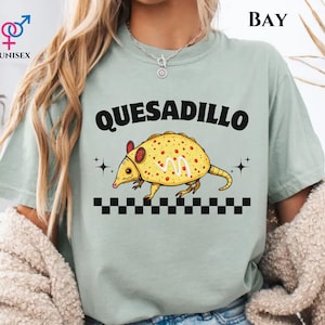 May include: A light green unisex t-shirt with the word "QUESADILLO" in black block letters above a cartoon armadillo-quesadilla hybrid. The armadillo is yellow with red spots and a checkered pattern at the bottom.
