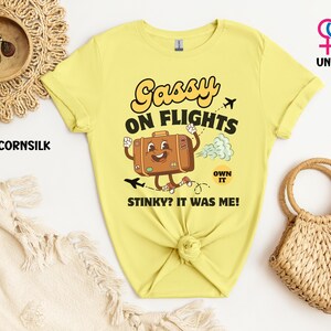 May include: A light yellow unisex t-shirt with the text "Sassy on Flights" and a cartoon suitcase graphic. The shirt also has the text "Stinky? It was me!" and "Own it".