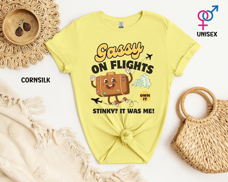 May include: A light yellow unisex t-shirt with the text "Sassy on Flights" and a cartoon suitcase graphic. The shirt also has the text "Stinky? It was me!" and "Own it".