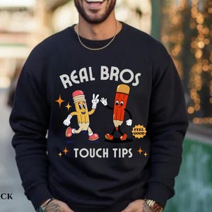 May include: Black sweatshirt with a graphic of two cartoon pencils, one yellow and one red, with the text "Real Bros Touch Tips" and "Feel Good" on a white circle.