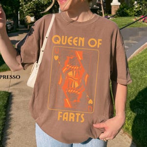 May include: Espresso-colored t-shirt featuring a playing card design with the words "QUEEN OF FARTS" in orange. The card depicts a stylized queen figure in orange. The shirt is a casual, oversized fit.