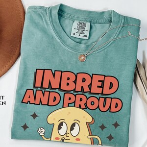 May include: A light green t-shirt with the text "Inbred and Proud" in red and white letters. A cartoon image of a slice of bread with eyes and a mouth is below the text.