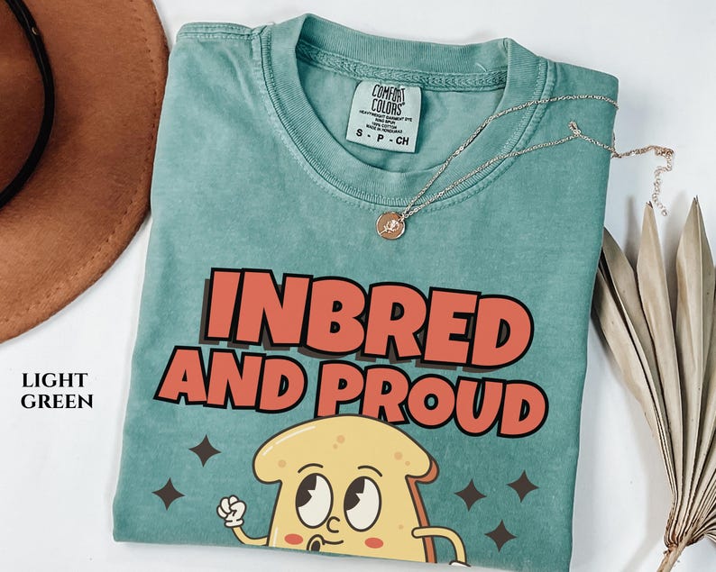 May include: A light green t-shirt with the text "Inbred and Proud" in red and white letters. A cartoon image of a slice of bread with eyes and a mouth is below the text.