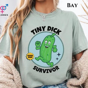May include: A light green unisex t-shirt featuring a cartoon pickle character with the text "TINY DICK SURVIVOR" inside a blue circle. The shirt also has the text "CERTIFIED KOSHER" on a yellow badge.