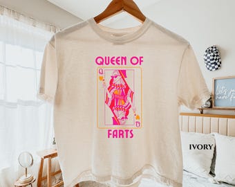 May include: Ivory-colored t-shirt with a graphic of a playing card featuring a woman in sunglasses. The text "QUEEN OF FARTS" is above and below the card image. The shirt is hanging on a wooden hanger.