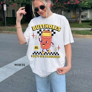 May include: White t-shirt with a graphic design featuring the text "BUTTHOLES DON'T GET PREGNANT". The design includes a cartoon illustration of a jar with a face, arms, and legs, set against a checkered background.