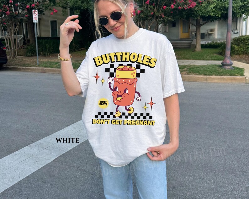 May include: White t-shirt with a graphic design featuring the text "BUTTHOLES DON'T GET PREGNANT". The design includes a cartoon illustration of a jar with a face, arms, and legs, set against a checkered background.