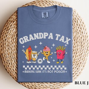 May include: Blue jean t-shirt with the words "GRANDPA TAX" in white, along with cartoon food characters. The shirt features a hot dog, coffee cup, cupcake, and french fries. The phrase "MAKING SURE IT'S NOT POISON" is also printed on the shirt.