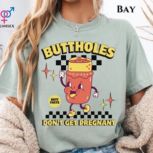 May include: A light green unisex t-shirt with the text "BUTTHOLES DON'T GET PREGNANT" in yellow and black. The shirt features a cartoon illustration of a jar with a face and arms.