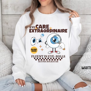 May include: White sweatshirt with a graphic of a cartoon eye wearing glasses and holding a magnifying glass. The text on the shirt reads "Eye Care Extraordinaire", "Blink Boss", "Fluent in Eye-Rolls", and "Retina Rockstar".