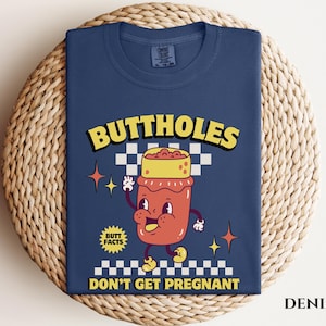 May include: Denim blue t-shirt with a cartoon graphic of a jar with a face, arms, and legs. The text reads "BUTTHOLES" above the jar and "DON'T GET PREGNANT" below. The shirt also has the text "BUTT FACTS".