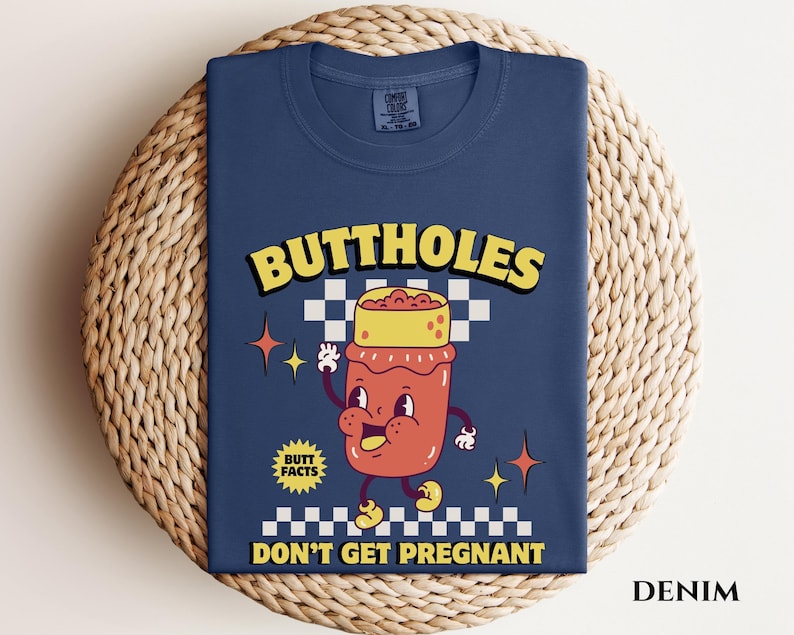 May include: Denim blue t-shirt with a cartoon graphic of a jar with a face, arms, and legs. The text reads "BUTTHOLES" above the jar and "DON'T GET PREGNANT" below. The shirt also has the text "BUTT FACTS".