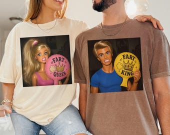 May include: A cream-colored t-shirt featuring a graphic of a blonde doll holding a pink balloon with the text FART QUEEN and a brown t-shirt featuring a graphic of a blonde doll holding a yellow balloon with the text FART KING.