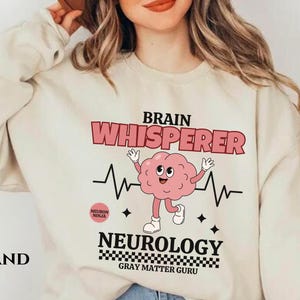 May include: A beige sweatshirt with a graphic design featuring a cartoon brain with a heartbeat and the text "Brain Whisperer Neurology Gray Matter Guru".