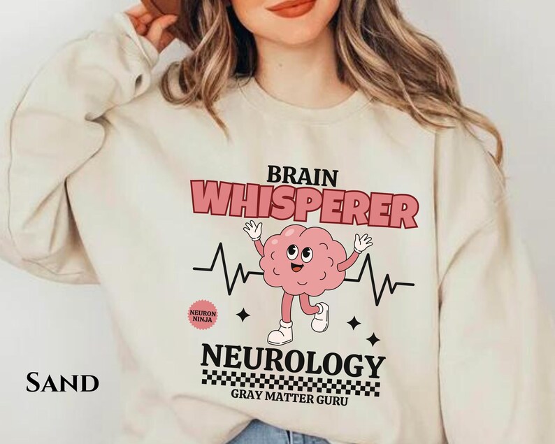 May include: A beige sweatshirt with a graphic design featuring a cartoon brain with a heartbeat and the text "Brain Whisperer Neurology Gray Matter Guru".