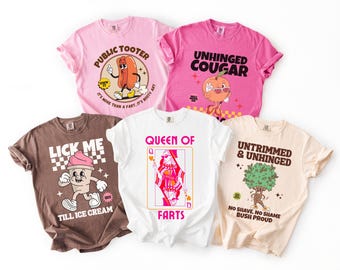 May include: Five colorful t-shirts with humorous cartoon graphics. Designs include a hot dog, ice cream, a playing card, a peach, and a bush. The shirts are in shades of pink, brown, and white, with various text elements.
