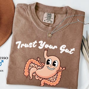 May include: Espresso-colored unisex t-shirt with the text "Trust Your Gut" and a cartoon stomach illustration. The shirt has a crew neck and short sleeves. A silver necklace with a pendant is draped across the shirt.