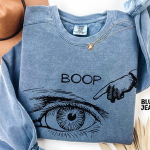 May include: Blue jean-colored sweatshirt with the word "BOOP" and a graphic of an eye and a pointing finger. The sweatshirt has a crew neck and long sleeves. A gold necklace with a pendant is draped on the sweatshirt.
