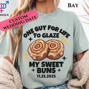 May include: A light green unisex t-shirt with the text "ONE GUY FOR LIFE TO GLAZE MY SWEET BUNS" and the date "11.25.2025". The shirt features a graphic of two cinnamon rolls. A pink banner reads "CUSTOM WEDDING DATE".