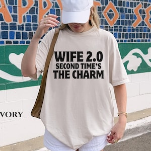 May include: Ivory-colored t-shirt with the text "WIFE 2.0 SECOND TIME'S THE CHARM" in bold black letters. The shirt is a relaxed fit. The word "IVORY" is printed in black at the bottom left of the shirt.
