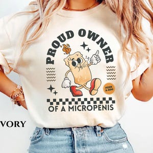 K&ouml;nnte beinhalten: Ein wei&szlig;es T-Shirt mit dem Text "PROUD OWNER OF A MICROPENIS" in schwarzer Schrift. Der Text ist von einem schwarz-wei&szlig;en Karomuster umgeben. Ein Cartoon-Kerze mit einem Gesicht und ein schwarz-wei&szlig;er "PEEK A-BOO" Aufkleber sind ebenfalls auf dem T-Shirt.