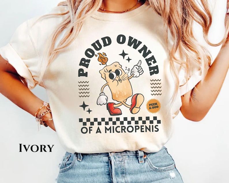 K&ouml;nnte beinhalten: Ein wei&szlig;es T-Shirt mit dem Text "PROUD OWNER OF A MICROPENIS" in schwarzer Schrift. Der Text ist von einem schwarz-wei&szlig;en Karomuster umgeben. Ein Cartoon-Kerze mit einem Gesicht und ein schwarz-wei&szlig;er "PEEK A-BOO" Aufkleber sind ebenfalls auf dem T-Shirt.