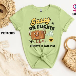 May include: A pistachio green unisex t-shirt with the text "Gassy on Flights" and a cartoon suitcase character. The suitcase is brown with a smiling face and is emitting a cloud of gas. The text "Stinky? It was me!" is also on the shirt.