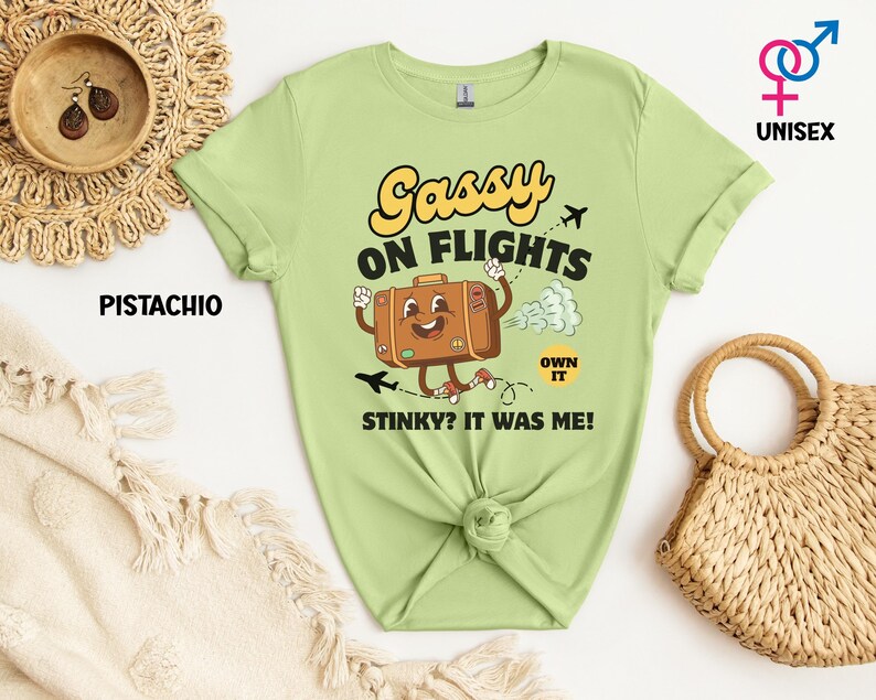 May include: A pistachio green unisex t-shirt with the text "Gassy on Flights" and a cartoon suitcase character. The suitcase is brown with a smiling face and is emitting a cloud of gas. The text "Stinky? It was me!" is also on the shirt.