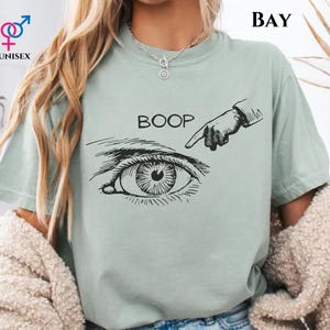 May include: A light green unisex t-shirt with a black graphic of an eye being poked by a finger, with the word "BOOP" above it. The shirt also has a gender symbol and the word "UNISEX" in the upper left corner. The word "BAY" is in the upper right corner.