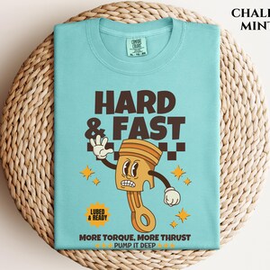 Puede incluir: Una camiseta azul turquesa con una ilustraci&oacute;n de dibujos animados de un pist&oacute;n con una cara. El texto "HARD & FAST" est&aacute; por encima del pist&oacute;n y "MORE TORQUE, MORE THRUST PUMP IT DEEP" est&aacute; por debajo del pist&oacute;n. La camiseta est&aacute; sobre una superficie de mimbre tejido.