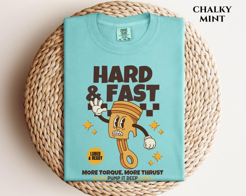 Puede incluir: Una camiseta azul turquesa con una ilustraci&oacute;n de dibujos animados de un pist&oacute;n con una cara. El texto "HARD & FAST" est&aacute; por encima del pist&oacute;n y "MORE TORQUE, MORE THRUST PUMP IT DEEP" est&aacute; por debajo del pist&oacute;n. La camiseta est&aacute; sobre una superficie de mimbre tejido.