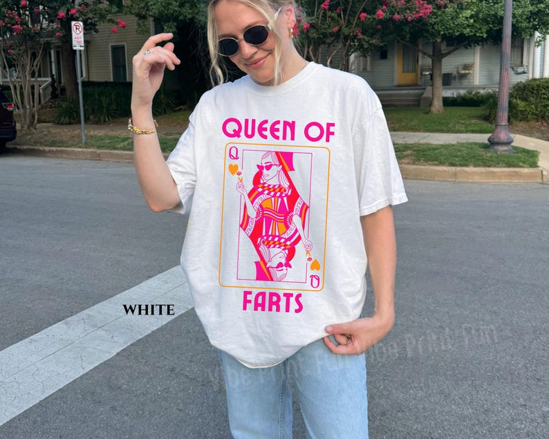 May include: White t-shirt with a graphic of a playing card featuring a queen wearing sunglasses, with the text "QUEEN OF FARTS" in pink. The shirt is a casual style, suitable for everyday wear.