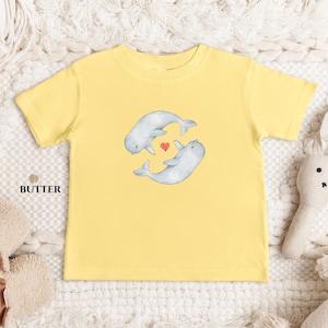 May include: A yellow t-shirt with a watercolour illustration of two beluga whales and a red heart. The shirt is made of soft material and is suitable for children. The word "BUTTER" is printed on the shirt.