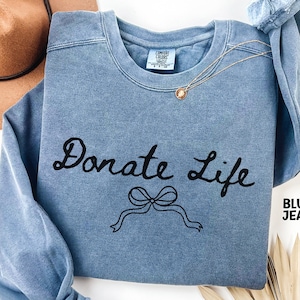 May include: Blue jean colored sweatshirt with the words "Donate Life" in black script, along with a black bow graphic. A gold necklace with a small pendant is draped across the sweatshirt. The tag reads "Comfort Colors".