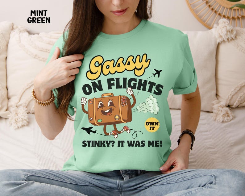 May include: Mint green t-shirt with the text "Gassy on Flights" and a cartoon suitcase character. The suitcase is brown with a smiling face and is running. The shirt also has the text "Stinky? It was me!"