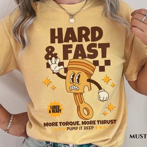 Puede incluir: Una camiseta amarilla mostaza con una ilustraci&oacute;n de dibujos animados de un pist&oacute;n con una cara. El texto "HARD & FAST" est&aacute; por encima del pist&oacute;n y "MORE TORQUE, MORE THRUST PUMP IT DEEP***" est&aacute; debajo del pist&oacute;n. El texto "LUBED & READY" est&aacute; en un peque&ntilde;o rect&aacute;ngulo por encima del pist&oacute;n.