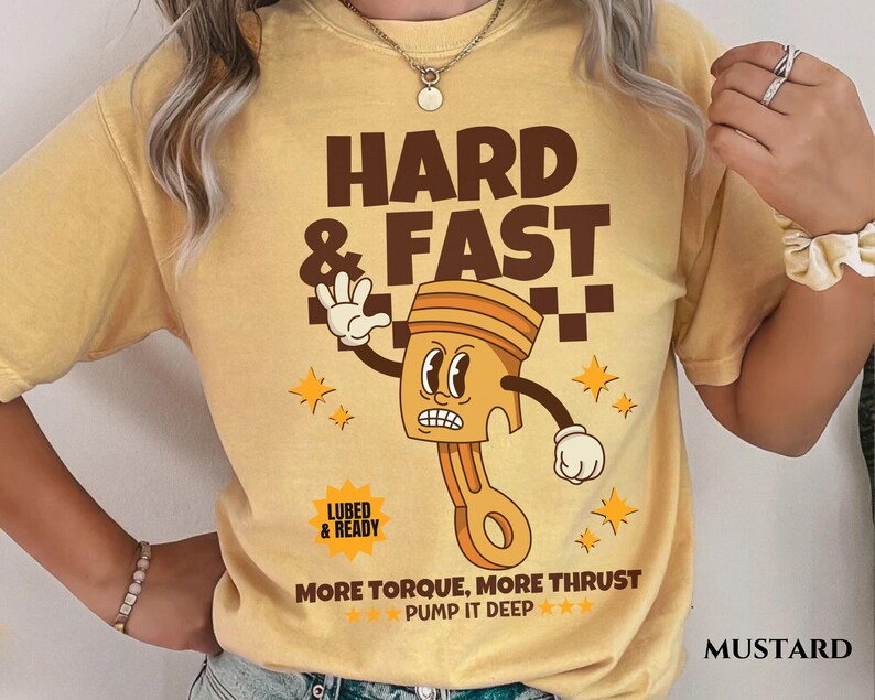Puede incluir: Una camiseta amarilla mostaza con una ilustraci&oacute;n de dibujos animados de un pist&oacute;n con una cara. El texto "HARD & FAST" est&aacute; por encima del pist&oacute;n y "MORE TORQUE, MORE THRUST PUMP IT DEEP***" est&aacute; debajo del pist&oacute;n. El texto "LUBED & READY" est&aacute; en un peque&ntilde;o rect&aacute;ngulo por encima del pist&oacute;n.